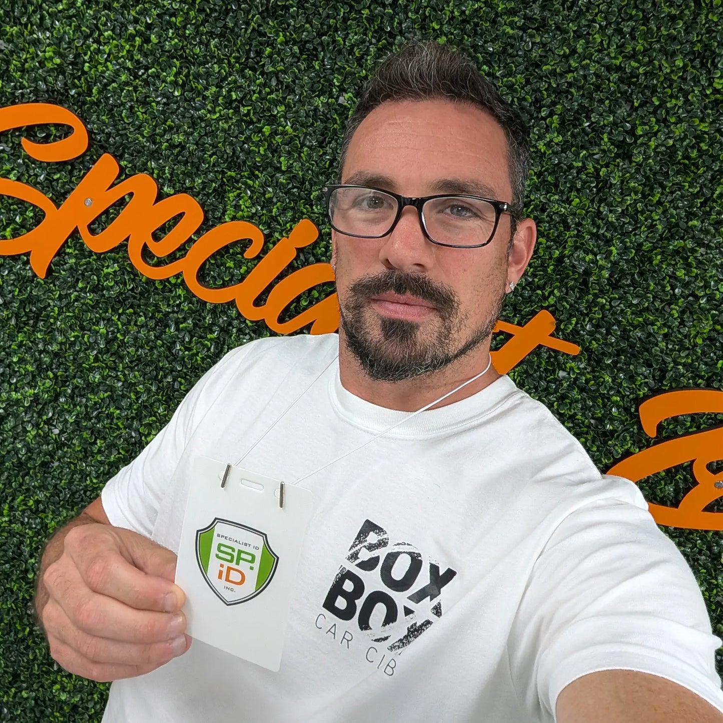 A man with glasses and a beard holds up an SPiD badge attached to an Elastic No-Twist Neck Cord for ID Event Badge Holders (2140-15XX Series) while standing in front of a green wall with orange cursive writing.
