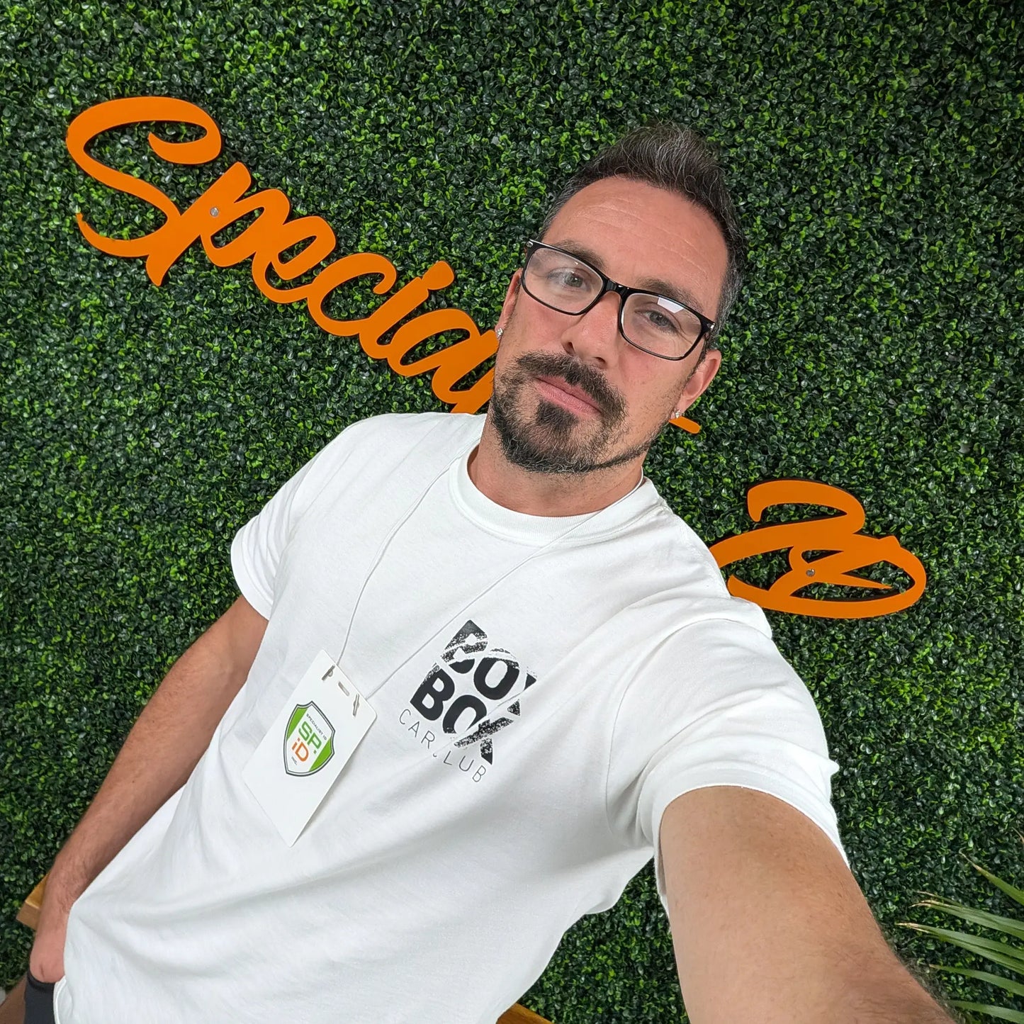 A man with glasses and a beard takes a selfie in front of a leafy wall with orange cursive text. He wears a white t-shirt and an event badge on the Elastic No-Twist Neck Cord for ID Badge Holders (2140-15XX Series) for a casual yet official style.