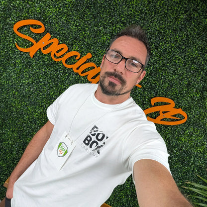 A man with glasses and a beard takes a selfie in front of a leafy wall with orange cursive text. He wears a white t-shirt and an event badge on the Elastic No-Twist Neck Cord for ID Badge Holders (2140-15XX Series) for a casual yet official style.