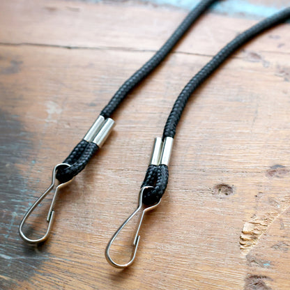 Close up of two black braided rope lanyards with metal hooks on a wooden surface. Sold by Specialist ID.