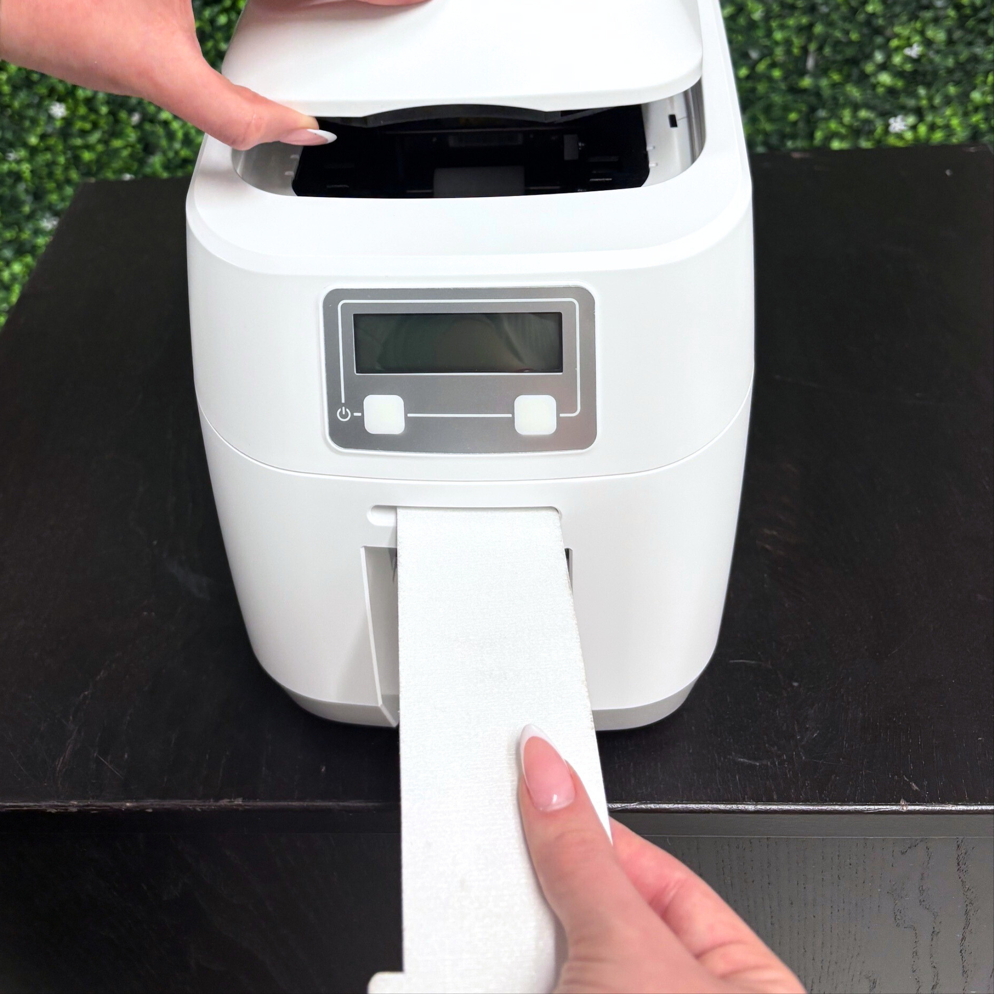 A person loads white label paper into a desktop label printer on a black table with a leafy background, demonstrating easy maintenance using the MAGICARD 3633-0053 Printer Cleaning Kit with Alcohol Cards & Printhead Pen.
