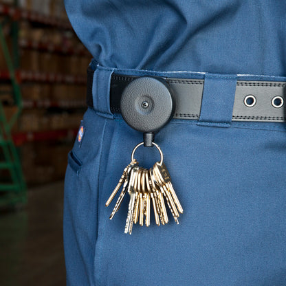 Close-up of a person in blue uniform pants with a black belt, holding a keyring with multiple keys attached to a Key-Bak #485B-HDK Self Retracting Key Reel. The background shows shelves and storage crates in a warehouse setting, indicating security enforcement.