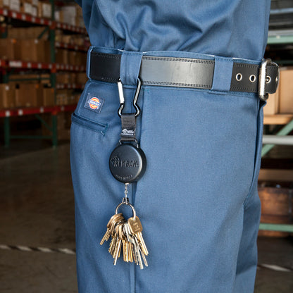 A person in blue work pants with a black belt is shown, featuring a Key-Bak SecureIt Super Duty Gear Reel #488B-SDK that holds multiple keys. Shelves and boxes are visible in the background.
