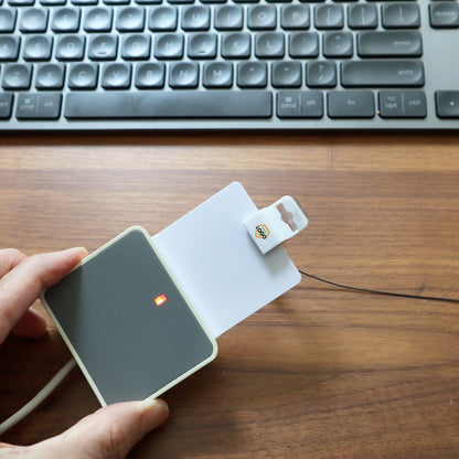 A person is using a Custom Printed Gripper 30 Card Clamp (5710-305X) from Brand it! to verify a small white card with a chip near a keyboard on a wooden desk. Sold by Specialist ID.