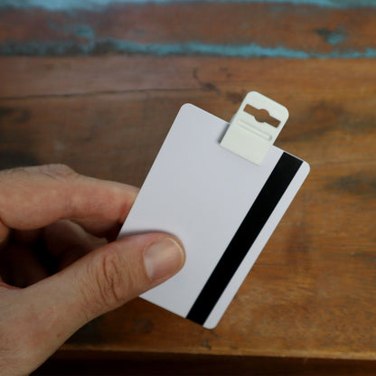 A hand holds a white card equipped with a magnetic stripe, secured by the Gripper 30 Card Clamp (P/N 5710-3058 White), featuring a plastic clip, all resting on a wooden surface. Sold by Specialist ID.