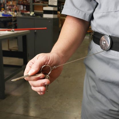 A person wearing a gray uniform holds keys attached to a Key-Bak #5 Self Retracting Key Badge Reel clipped onto their belt in a workspace.