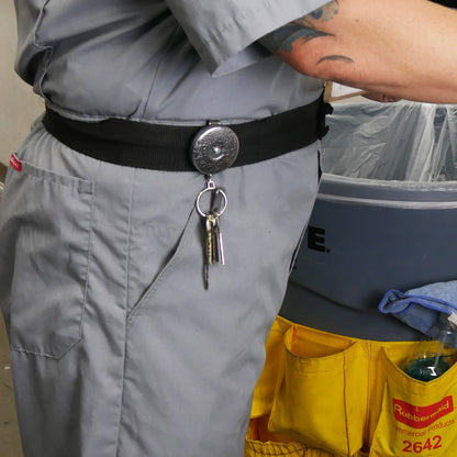 A person in a grey uniform is standing next to a trash can, with keys on a Key-Bak #5 Self Retracting Key Badge Reel clipped to their belt and a caddy with cleaning supplies attached to the trash can.