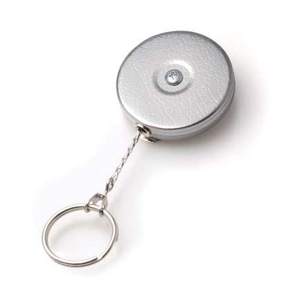 A round, silver Key-Bak #5 Self Retracting Key Badge Reel with a textured pattern and a stainless steel chain. The surface features a screw in the center, and the durable Kevlar cord ensures reliability.