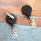 Two Premium No Twist Badge Reels with Swivel Spring Clips (teardrop shape, 609-I-BLK) in black rest on a wooden surface—one face up, one face down. Their retractable cord lets you display your ID easily without twisting.