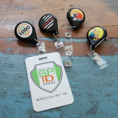 Four Custom No Twist Teardrop Badge Reels with swivel spring clips (1" imprint area) and a white SPIC INC name badge are displayed on a rustic wooden surface, highlighting their unique design and eye-catching presentation.