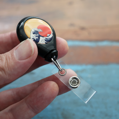 A hand holds the Custom No Twist 1 Inch Imprint Area Badge Reel with Swivel Spring Clip - Teardrop Shape, featuring a wave and red sun design, attached to a clear plastic strap with snap button.