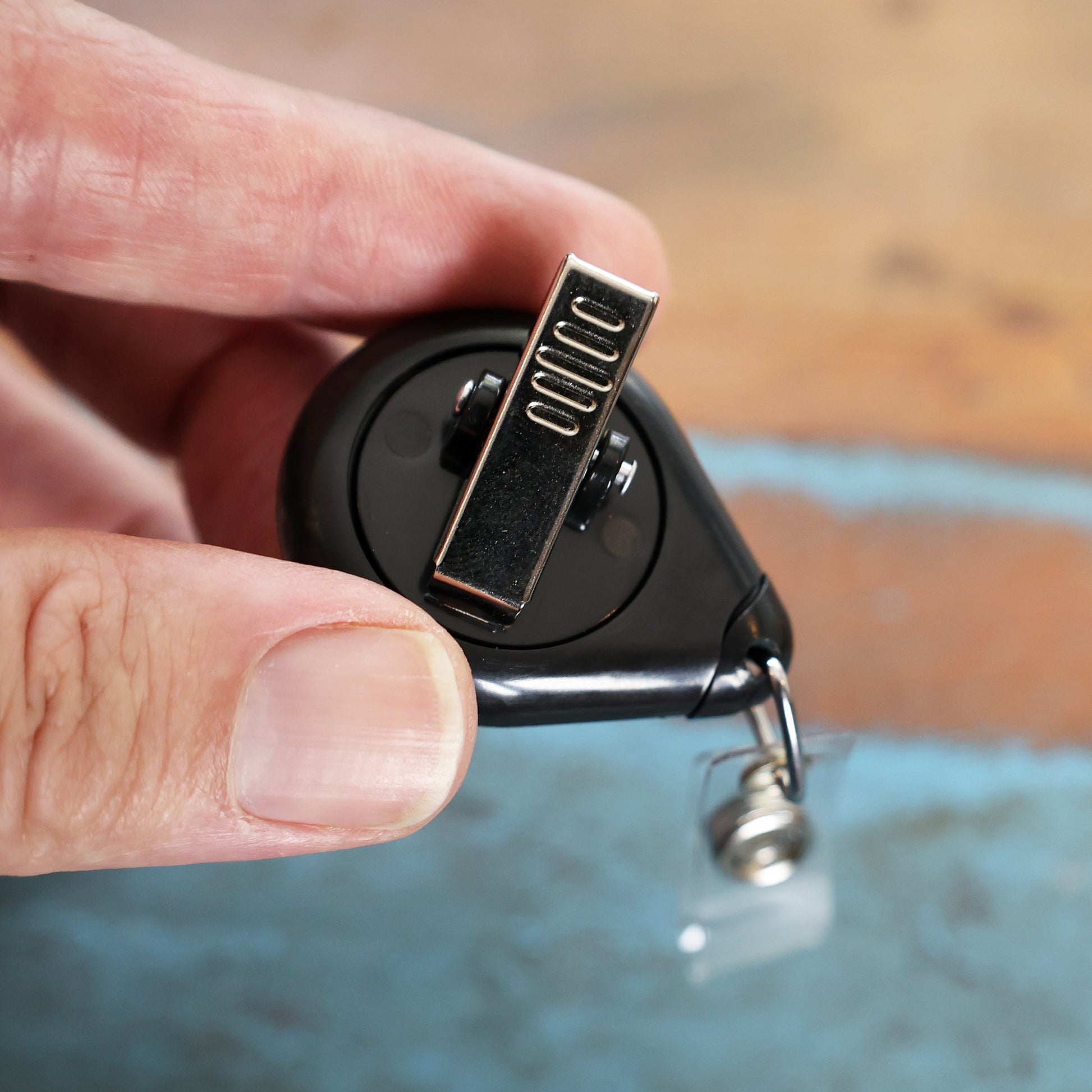 A hand holds a Premium No Twist Badge Reel with Swivel Spring Clip, Teardrop Shape (609-I-BLK), featuring a black retractable reel and transparent plastic strap.