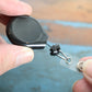 A hand holds the Premium No Twist Badge Reel with Swivel Spring Clip – Teardrop Shape (609-I-BLK), featuring a black retractable cord and key ring attachment.