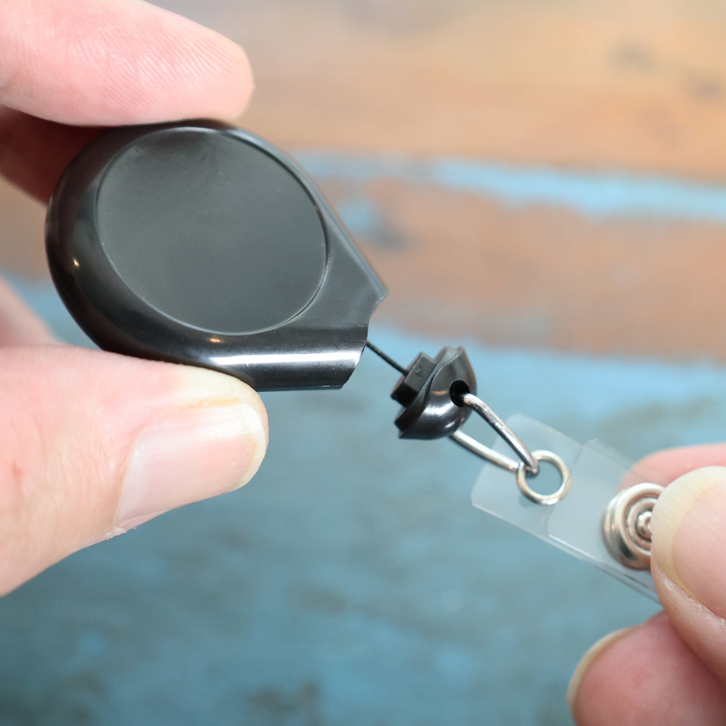 A hand holds the Premium No Twist Badge Reel with Swivel Spring Clip – Teardrop Shape (609-I-BLK), featuring a black retractable cord and key ring attachment.
