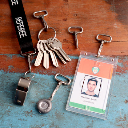 A set of keys on a lanyard labeled "REFEREE," featuring 1 3/4" Non Swivel J Hook with Rectangular End - DIY Lanyard and Craft Accessories (6920-2400), a whistle, three key clips, a badge with the name "Raphael Smith" listed under "Operations," and a held metal object on a blue and brown surface.