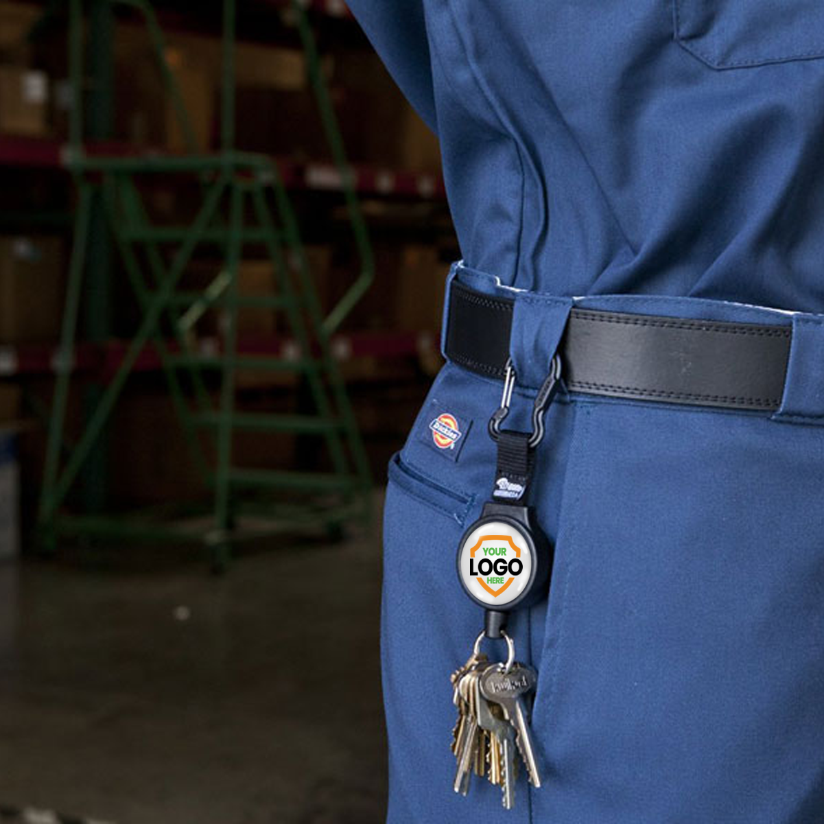 Close-up of a person wearing a blue uniform with a key ring attached to the belt by a Customizable Key-Bak 6C Carabiner Badge Reel - Retractable Keychain Reel with 36" Cord and Easy Logo Upload, promoting brand awareness. Shelves with boxes are visible in the background.
