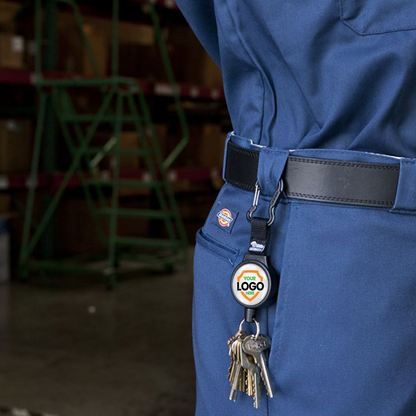 Close-up of a person wearing a blue uniform with a key ring attached to the belt by a Customizable Key-Bak 6C Carabiner Badge Reel - Retractable Keychain Reel with 36" Cord and Easy Logo Upload, promoting brand awareness. Shelves with boxes are visible in the background.