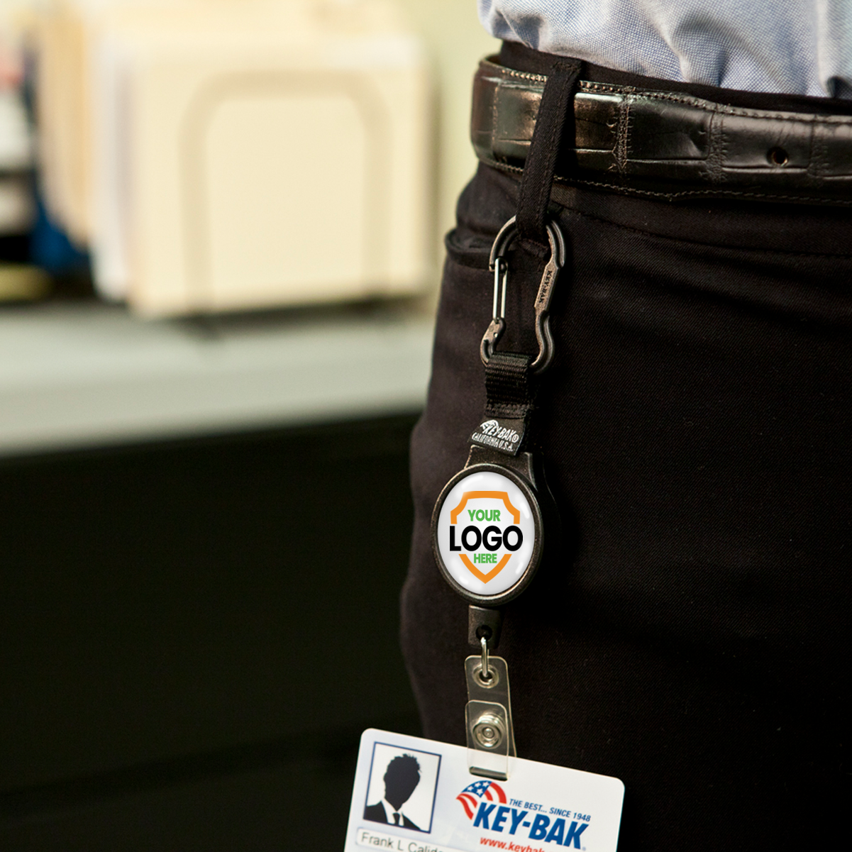 A person wearing a belt with a Customizable Carabiner Badge Reel Retractable Badge Holder with Vinyl Strap Clip (6CID) - Add Your Logo. The badge holder is clipped to the belt, with a card attached at the end.