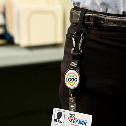 A person wearing a belt with a Customizable Carabiner Badge Reel Retractable Badge Holder with Vinyl Strap Clip (6CID) - Add Your Logo. The badge holder is clipped to the belt, with a card attached at the end.