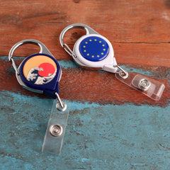 Two Custom Printed No Twist Carabiner Badge Reels - Upload Your Logo on a wooden surface. One showcases a wave and red sun design, while the other features the EU flag with yellow stars against a blue background.