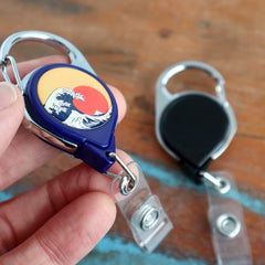 A person's hand holding a retractable badge holder featuring a design inspired by The Great Wave off Kanagawa, with another plain black Custom Printed No Twist Carabiner Badge Reel - Upload Your Logo on the table in the background.