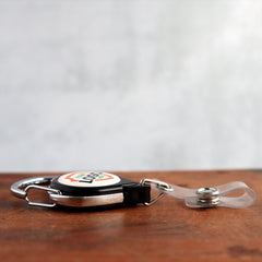 A Custom Printed No Twist Carabiner Badge Reel - Upload Your Logo with a keyring and transparent plastic strap is placed on a wooden surface. The custom badge reel has a round, white center featuring a logo.