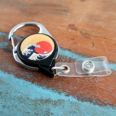 A Custom Printed No Twist Carabiner Badge Reel - Upload Your Logo with a retractable clip features a round design depicting a stylized wave and red sun on a wooden surface. Sold by Specialist ID.