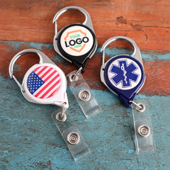 Three Custom Printed No Twist Carabiner Badge Reels - Upload Your Logo, featuring different designs: one with an American flag, one with a medical symbol, and one with space for a personalized logo badge. They are on a wooden surface.