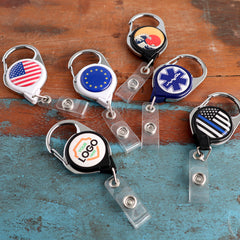 Six Custom Printed No Twist Carabiner Badge Reels - Upload Your Logo with different designs, including flags of the United States, European Union, a medical symbol, a custom logo personalized on custom badge reels, and a thin blue line flag, on a wooden surface.