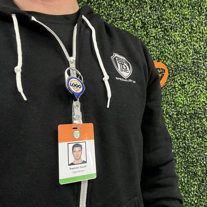 Person wearing a black hoodie with a visible ID badge, attached to a Translucent Custom Carabiner Badge Reel With No Twist Strap Clip (704-TR), showing a photo, the name "Raphael Smith," and the word "Operations." Background consists of green foliage.