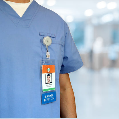A person in blue medical scrubs, wearing a NURSE PRACTITIONER BadgeBottom Badge Holder—a vertical ID badge sleeve with an integrated bottom role tag—stands against a blurred hospital backdrop.