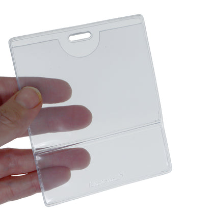 A hand holds a Premium Vinyl Horizontal BadgeBottom with Slot - Holder Only, transparent and rectangular, with a top slot for hanging. Sold by Specialist ID.