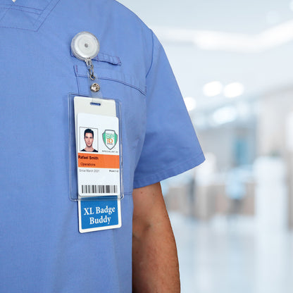 A person wearing a blue scrub outfit with a hospital ID badge featuring an "Extra Large Doctor Badge Buddy" tag enhances physician visibility. The background is a blurred clinical setting, emphasizing their role in healthcare identification.