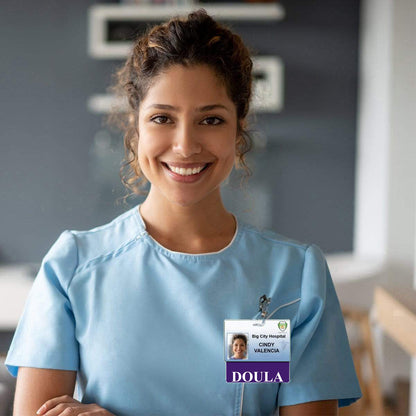 A smiling woman in light blue scrubs wears a hospital ID badge that reads "Doula - Cindy Valencia" in an indoor setting, her role recognition clearly marked by a DOULA Horizontal Badge Buddy with Purple Border.