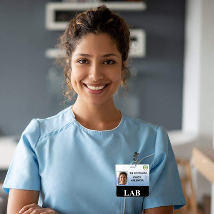 A woman in a blue medical uniform is smiling at the camera. She has curly hair and proudly wears her **LAB Badge Buddy Horizontal with Black Border - Badge Backer to Wear Behind Standard ID Card** that reads "Big City Hospital, Cindy Valencia, LAB.