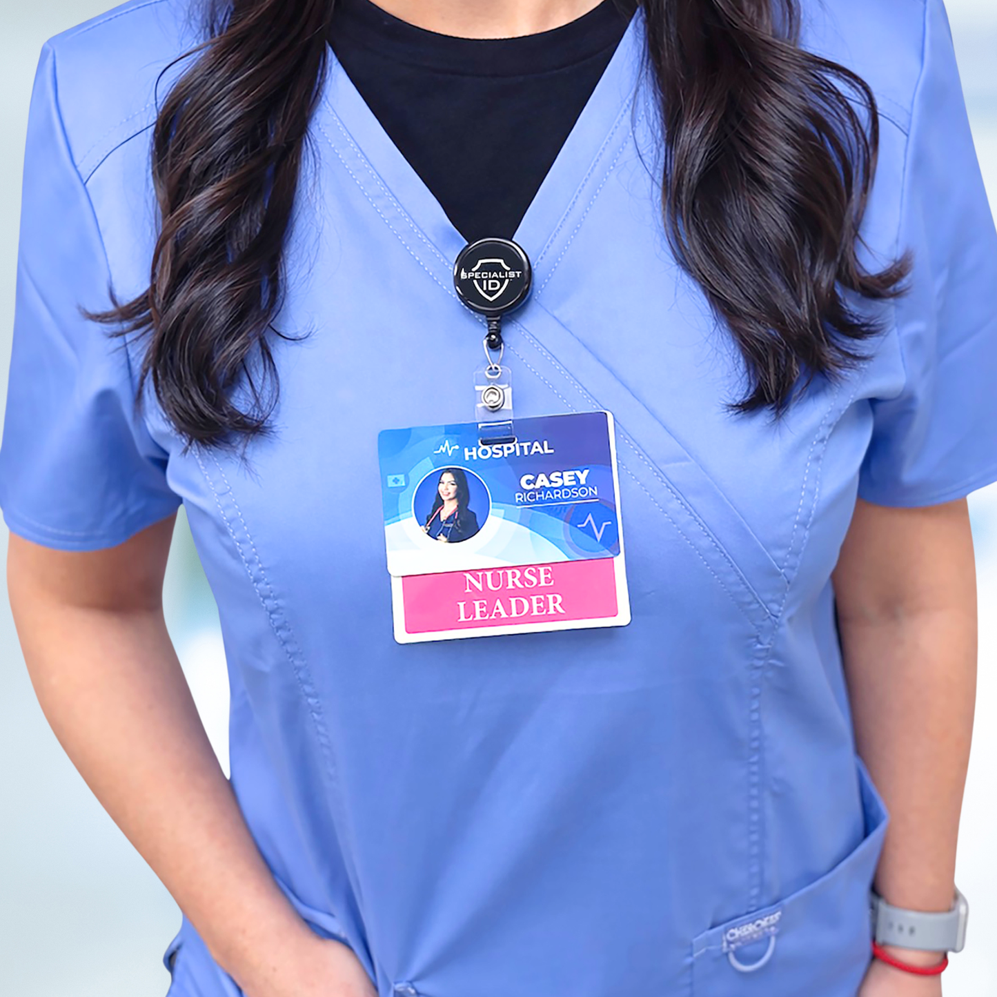 A person in blue scrubs stands indoors wearing a Nurse Leader Badge Buddy - Horizontal Hospital ID Role Identifier with their hospital ID clipped on top.
