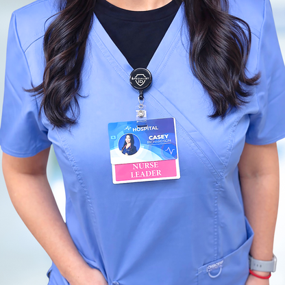 A person in blue scrubs stands indoors wearing a Nurse Leader Badge Buddy - Horizontal Hospital ID Role Identifier with their hospital ID clipped on top.