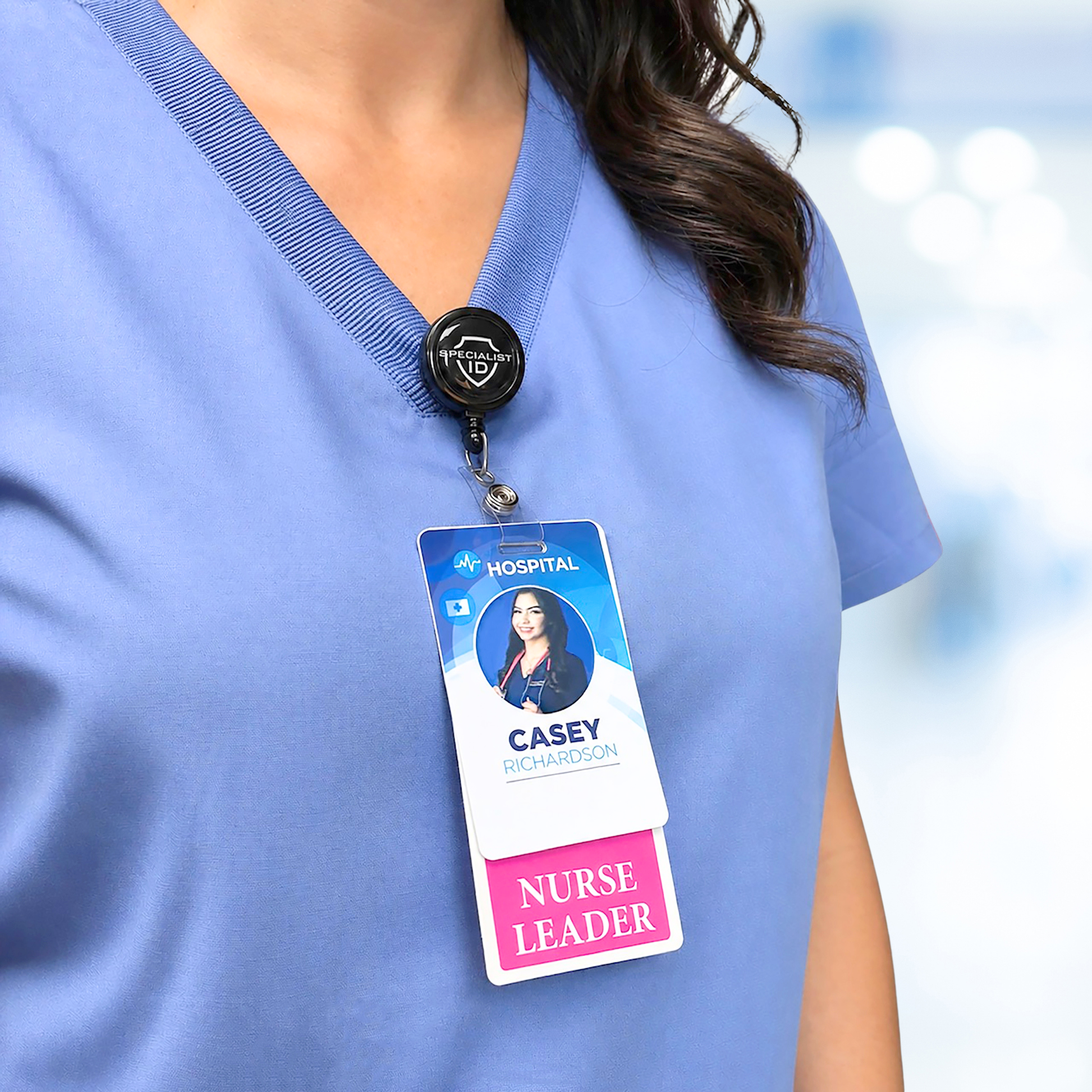A person in blue scrubs and a hospital ID featuring the durable "Nurse Leader Badge Buddy - Vertical Hospital ID Role Identifier" labeled “Casey Richardson,” stands indoors with a blurred background.