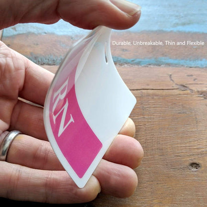 A hand holding a thin, flexible, and slightly curved plastic card with "RN" on a pink section and a green border. The LVN horizontal Badge Buddy with Green Border is described as durable, unbreakable, thin, and flexible.