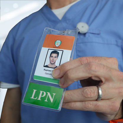 A person in blue scrubs, wearing a ring, holds an ID badge showing a photo, the name "Raphael," and the title "Operations." The badge features a green section labeled "LPN" and is secured by an LPN BadgeBottom Badge Holder & Badge Buddy IN ONE!! - Vertical ID Badge Sleeve with Bottom Role Tag for Nurses.