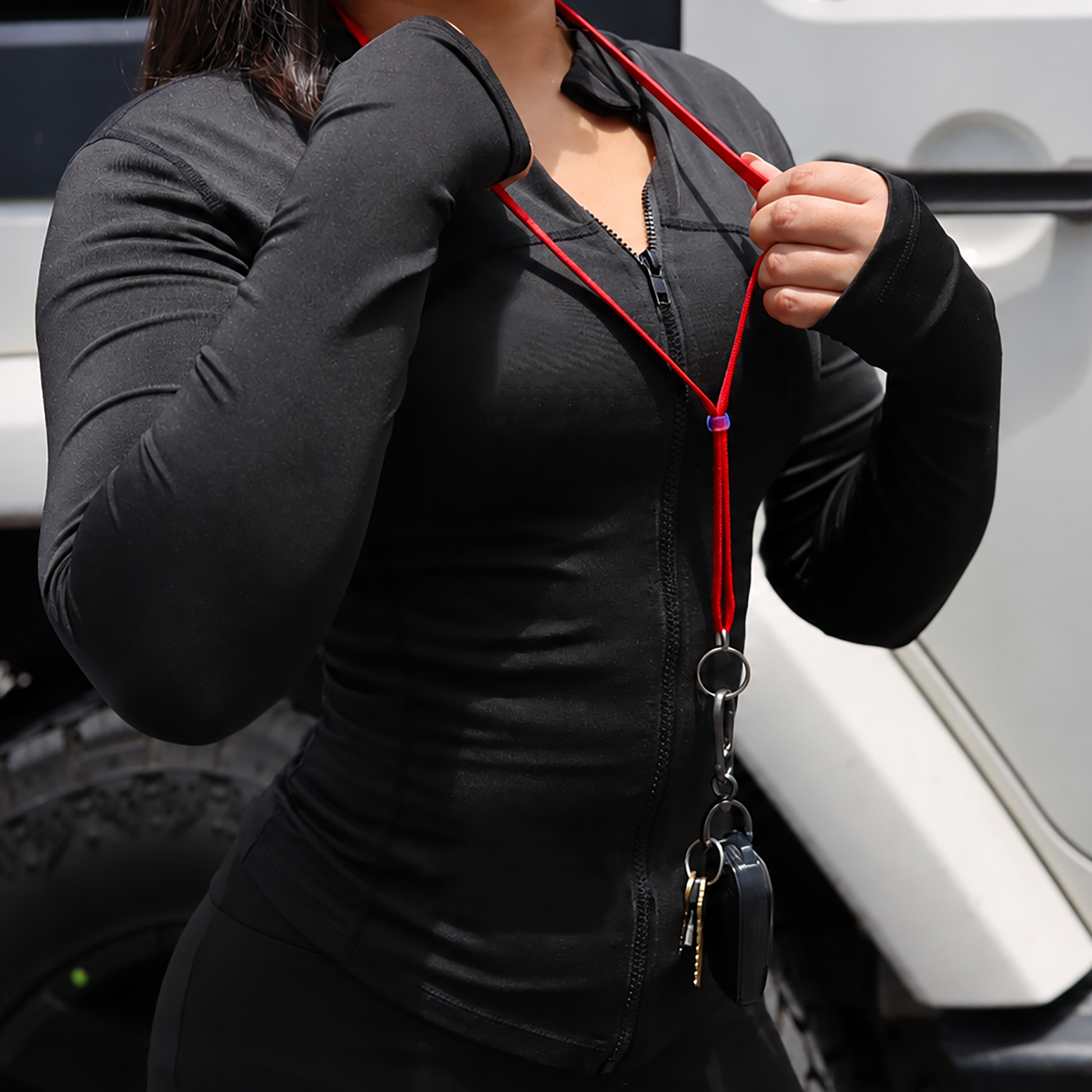 A close-up of a person wearing a fitted black zip-front long-sleeve top and matching black gloves, holding a red lanyard with a key fob and keys in front of a white vehicle.