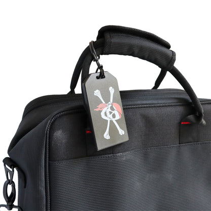 A close-up of a black travel bag with a skull and crossbones luggage tag attached to the handle via 6" Plastic Luggage Tag Loop Straps in Solid Colors - Red, Green, Black, Blue & White (2410-200X). The tag features a red scarf on the skull.