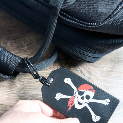A black luggage tag with a skull and crossed bones design is securely attached to a black bag using 6" Plastic Luggage Tag Loop Straps in Solid Colors - Red, Green, Black, Blue & White (2410-200X), making it a perfect accessory for all your travel bags.