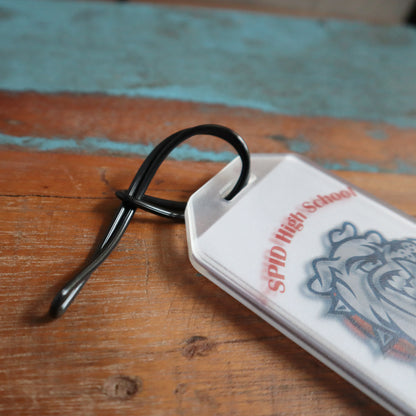 A white plastic keychain with a logo and the text "SPID High School" lies on a wooden surface, accompanied by 6" Plastic Luggage Tag Loop Straps in Solid Colors - Red, Green, Black, Blue & White (2410-200X). The keychain has a twisted black wire loop attached, perfect for securing it to travel bags.