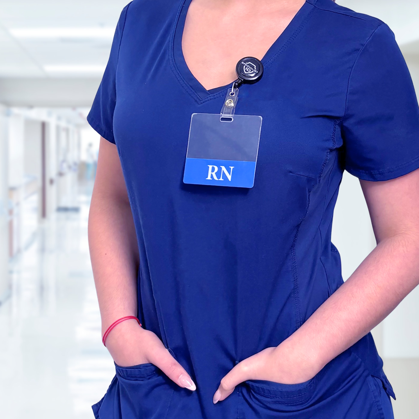 A person in blue scrubs with a nurse badge holder and a Clear RN Badge Buddy (horizontal, double-sided) stands in a hospital hallway with hands in pockets.