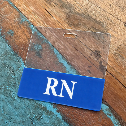 A Clear RN Badge Buddy - Horizontal ID Badge Backer for Nurses - Double Sided Print with a blue bottom, featuring the letters "RN" printed in white, serves as an ideal RN Badge Buddy on a wooden and blue surface.