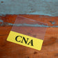 A rectangular Clear CNA Badge Buddy - Horizontal ID Badge Backer for Nursing Assistant - Double Sided Print with "CNA" printed in bold black letters on a yellow background lies on a wooden surface.