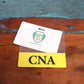 An identification card and its plastic holder rest on a textured wooden surface. The card, akin to a Clear CNA Badge Buddy - Horizontal ID Badge Backer for Nursing Assistant - Double Sided Print, features the acronym "CNA" in black on a yellow background.
