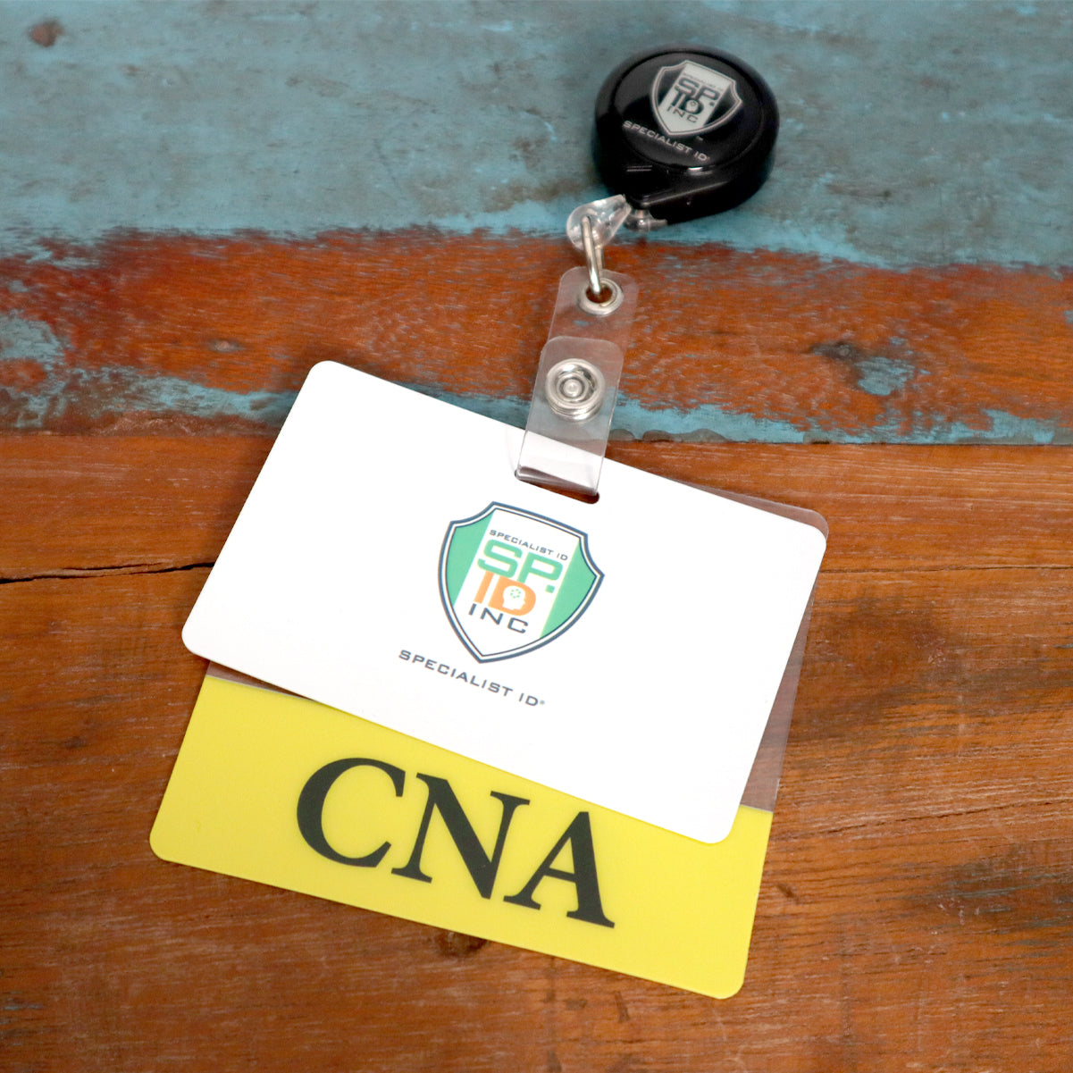 Close-up of a badge reel with two cards attached: one with the logo "SP inc" and another with "Clear CNA Badge Buddy - Horizontal ID Badge Backer for Nursing Assistant - Double Sided Print" in bold letters. The badges, perfect for nurse assistants, rest on a wooden surface.