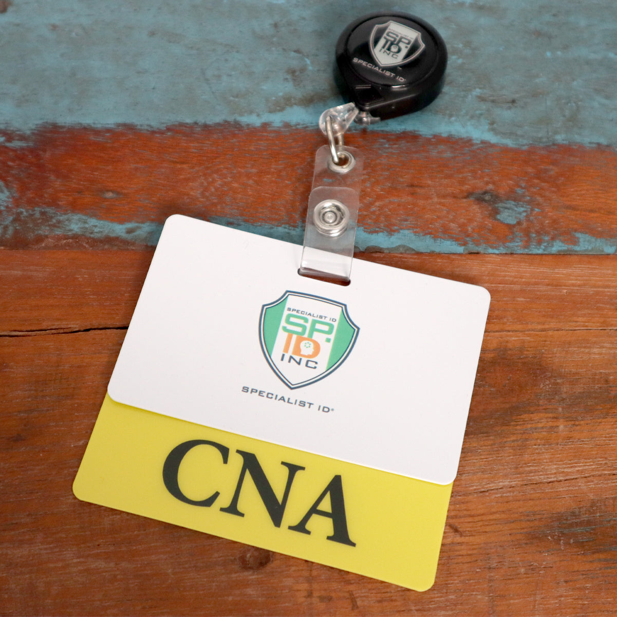 A name badge reel and holder rest on a wooden surface, featuring a "Specialist ID Inc." card and a yellow "CNA" card. The setup is complemented by a Clear CNA Badge Buddy - Horizontal ID Badge Backer for Nursing Assistant - Double Sided Print, ensuring the CNA Badge Buddy stays visible and secure.